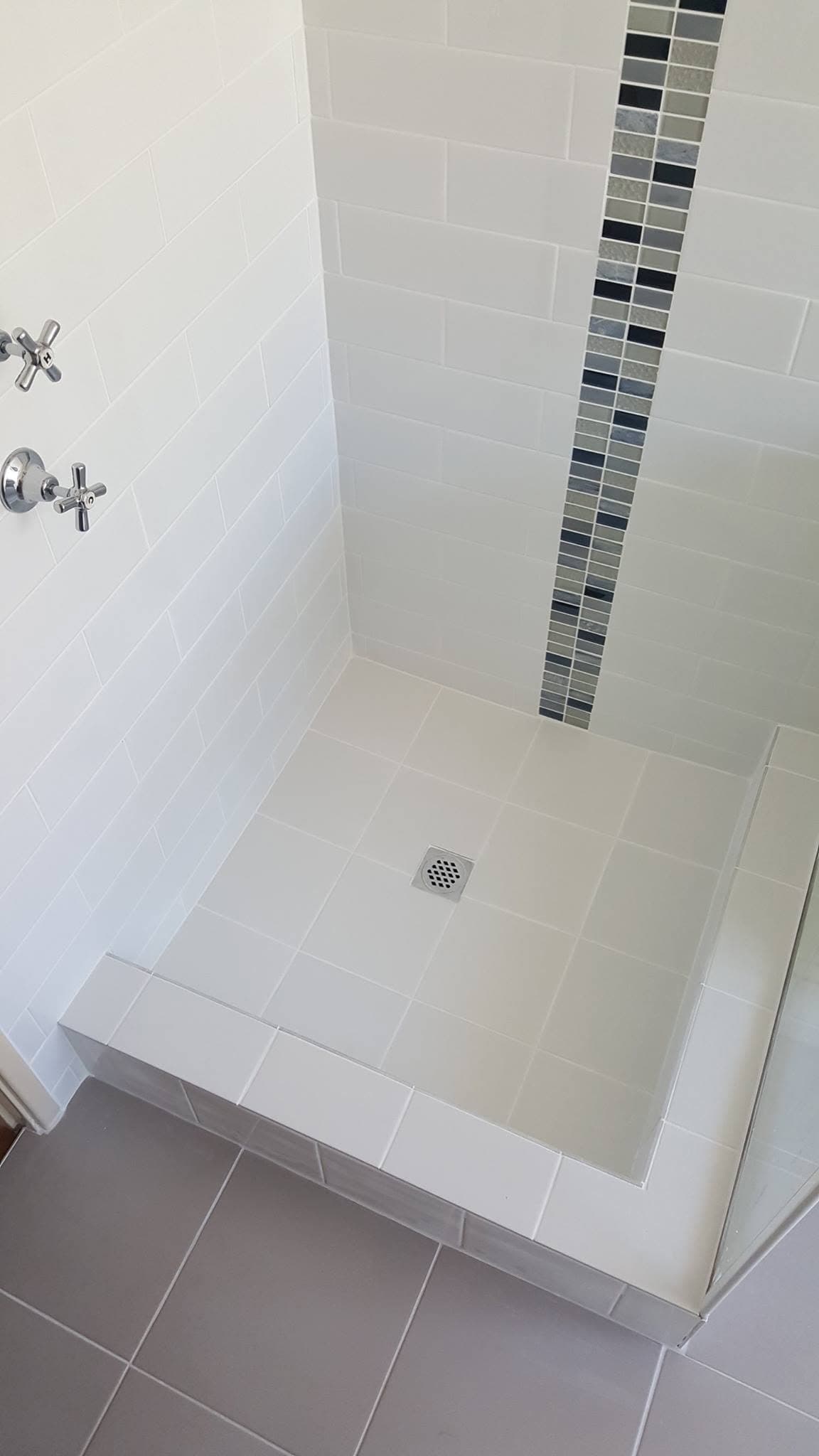 Tiled shower floor and hob with a vertical mosaic feature strip on the wall above, tapware visible