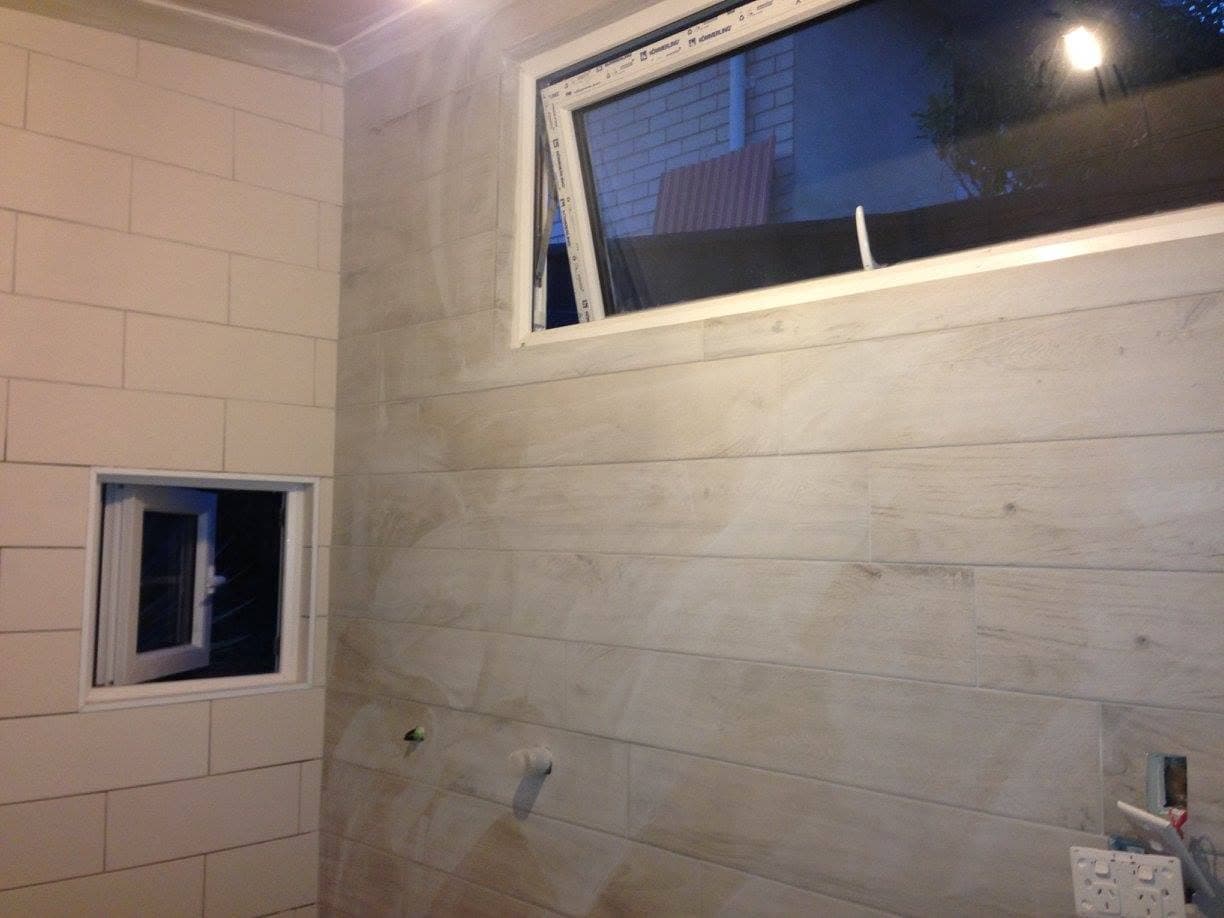 Wood-look plank tile being installed on a bathroom wall around two window openings, with plumbing rough-ins visible