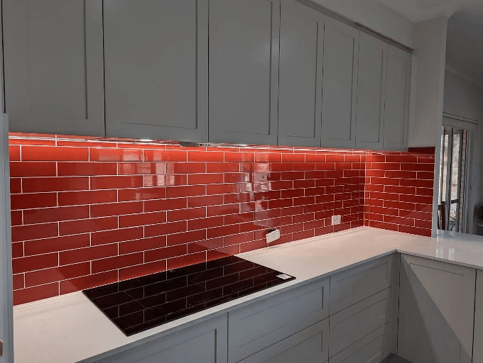 Red gloss subway tile kitchen splashback behind a black induction cooktop with grey cabinetry