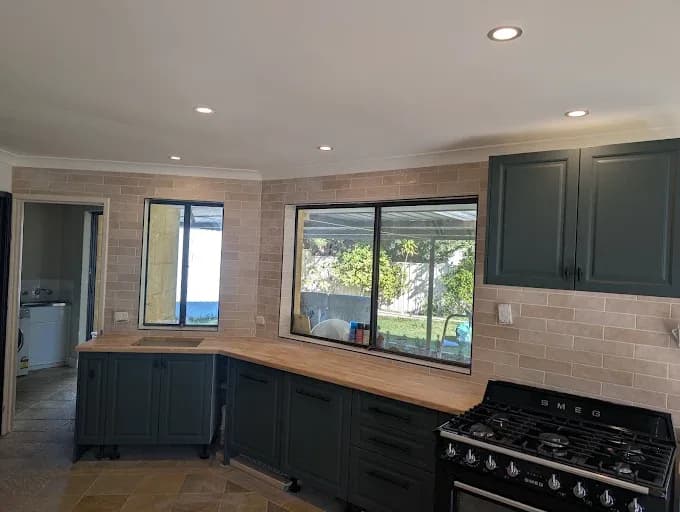 Kitchen with a sand-coloured brick-pattern tile splashback wrapping around windows, dark green cabinetry and a timber benchtop