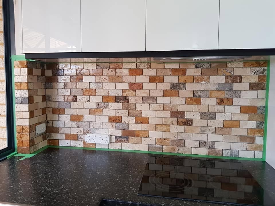 Multi-tone travertine brick-pattern tile kitchen splashback behind a black stone benchtop and induction cooktop
